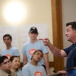 Camp instructor engaging with diverse group of children and teens in classroom.