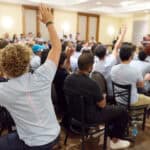 Audience members raising hands during a conference or seminar in a large room.