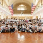 Large group of diverse children and adults in a spacious hall with American flags.