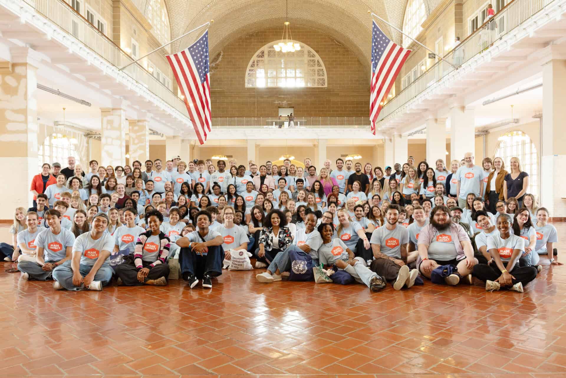 Large group of diverse children and adults in a spacious hall with American flags.