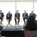 Panel of six men in suits speaking at a conference with audience in foreground.